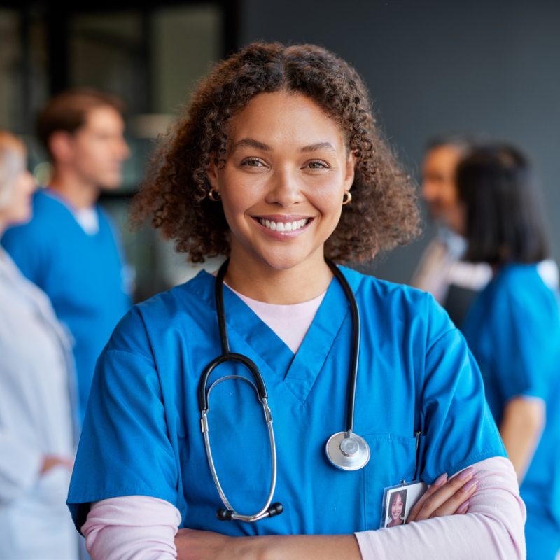 Confident happy woman nurse smiling with crossed arms with healthcare team in background. Multiethnic medical professional in scrub ready to assist patients. Portrait of reliable confident nurse standing proudly, surrounded by colleagues in a private clinic.